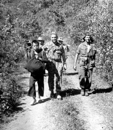 Trois hommes pris en photo dans une colonne en  progression :a gauche un porteur réquisitionné qui ne cache pas son mécontentement, au milieu le Lieutenant Roger PHILIPPON cigarette aux lèvres parait d' excellente humeur .Il est revètu d'une combinaison camouflées U.S. Sur sa tète ,visière relevée une casquette au camouflage US .A droite un Officier revètu de la tenue de saut kaki foncé (modèle 1947 Française )