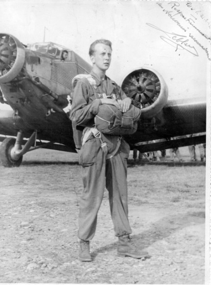 L'homme aux deux mains sur le ventral pose devant un JU 52 moteurs tournants On dirait qu'il est revètu d'une combinaison ..Le pilote observe l' embarquement des parachutistes partiellement visibles sous l' aile .