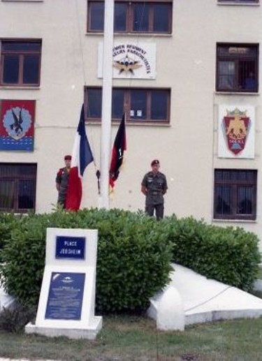 Prise de commandement conjointe alors que les couleurs Allemandes et Françaises sont prètes à être montées Sur la façade de la tour de commandement du 1° RCP les insignes du 1° RCP, de la 11° Division Parachutiste , du 144 Régiment d' Infanterie (réserve ).Au pied du mat des Couleurs la plaque d'inauguration de la Place JEBSHEIM  ( Colonel A.FAYETTE  au CAMP d' IDRON) "