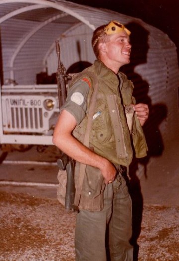 Un Caporal très souriant  galons métal peints en rouge sur les épaules ,se prépare à partir en mission. Armé d'un SIG 5,56 il semble porter une cartouchière toile de type TAP pour PM Mat 49. Au second plan la jeep 800 (celle du Chef de Section) dans son abri.